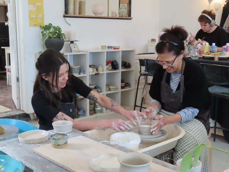 Two women are sitting at a pottery wheel, making a small clay pot